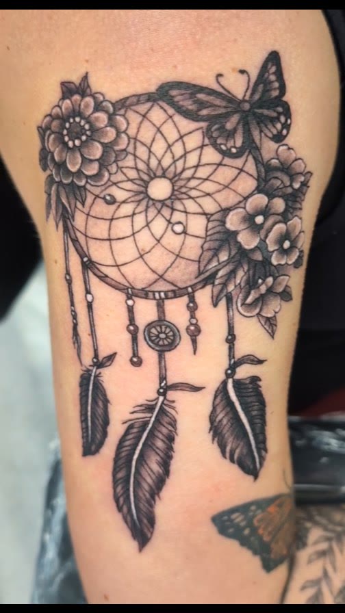 Dreamcatcher tattoo with flowers and feathers on an upper arm.